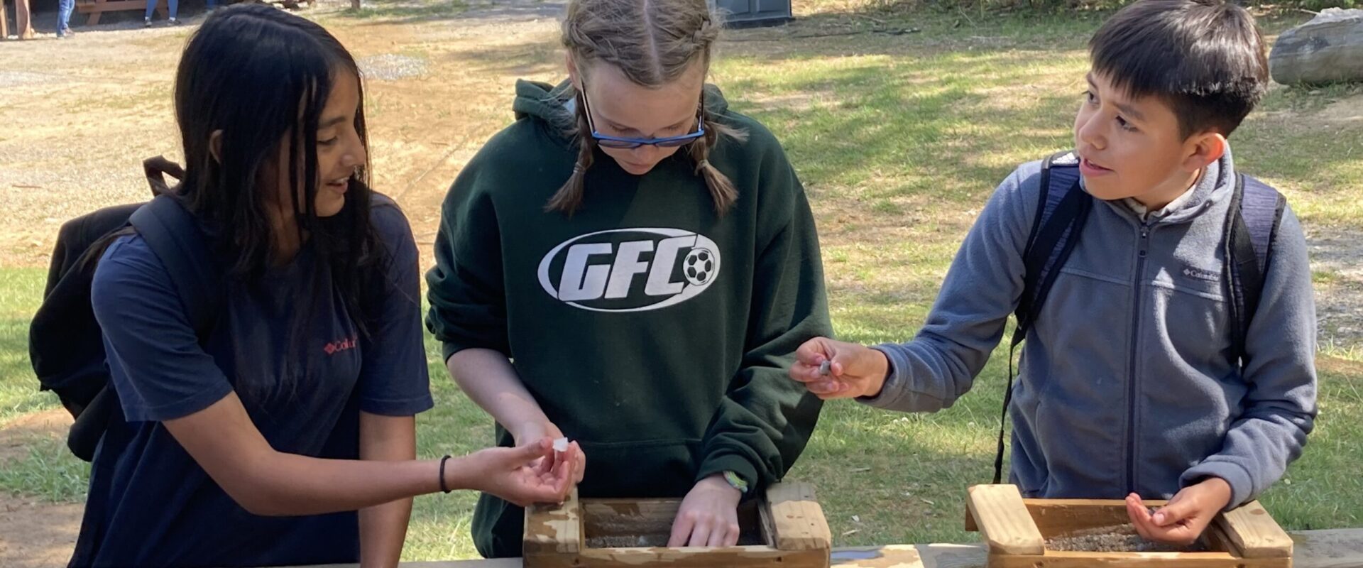 Three students pan for minerals at an outdoor trough